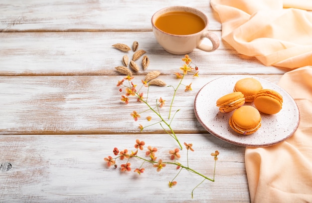 Orange Macarons oder Macaroons Kuchen mit Tasse Aprikosensaft auf einem weißen Holztisch und orange Leinen Textil. Seitenansicht, Kopierraum.