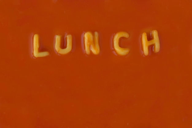 Foto orange pasta-buchstaben, die das wort mittagessen in roter tomatensauce schreiben, von oben betrachtet