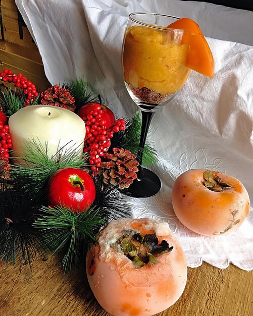 Orange Persimmon mit Eis bedeckt, umgeben von einem Nadelbaumzweig mit rotem Apfel und roten Beeren mit Kerzen auf der linken Seite auf einem hellen Hintergrund