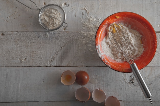 Orange Schüssel mit Mehl, Eiern und einem Schneebesen, um den Teig auf einem weißen Holzboden zu machen.