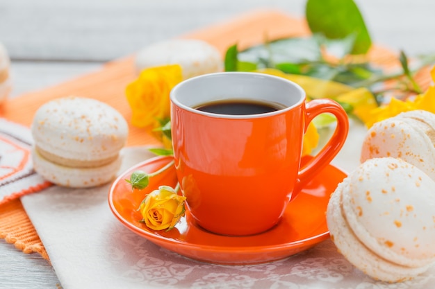Orange Tasse schwarzer Kaffee, gelbe Rosenblüten und süße französische Pastellmakronen auf weißem Holztisch