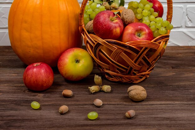 Orangefarbene Kürbisse mit Äpfeln und Birnen in einem Weidenkorb mit Trauben, Nüssen und Blättern auf einem braunen Holztisch vor einem weißen Backsteinmauerhintergrund Nahaufnahme Stillleben im Herbst