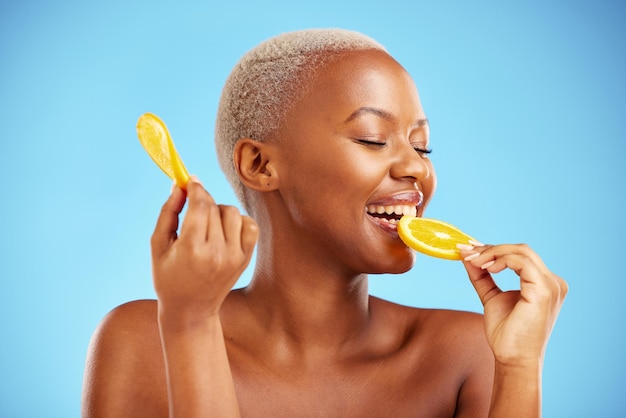 Orangefarbene Schönheit und Ernährung mit einer modellhaften schwarzen Frau im Studio auf blauem Hintergrund, die Früchte beißt. Lebensmittel, Hautpflege und Natur mit einer glücklichen jungen Frau, die einen Snack für Gesundheit oder Wohlbefinden isst