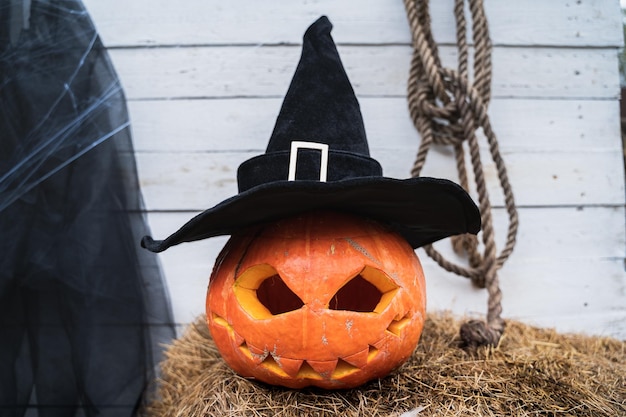 Orangefarbener Kürbis für Halloween in schwarzem Hexenzaubererhut Jackolantern mit unheimlich geschnitzten Augen Mund Heu Heuhaufen in der Nähe von Scheune Heimwerken Straßendekoration Unterhaltung für Kinder Horror