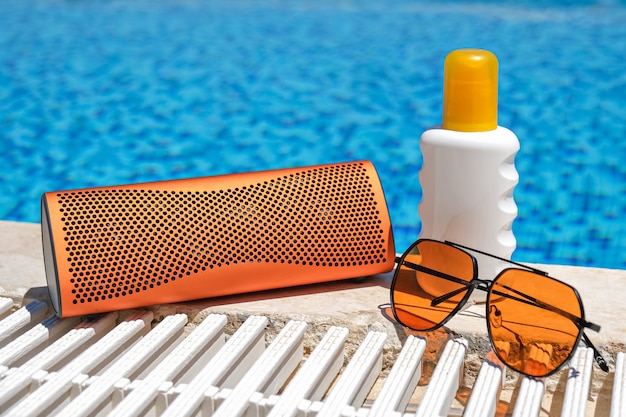 Orangefarbenes strandzubehör in der nähe des schwimmbades. sonnencreme, sonnenbrille, musik bluetooth-lautsprecher.