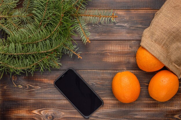 Orangen in einer Leinentasche auf einer Holzoberfläche nahe Tannenzweigen und schwarzen Smartphone Weihnachten