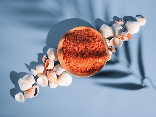 Foto orangenbadesalz in einer untertasse mit muscheln auf einem blauen hintergrund mit einem schatten von einer tropischen pflanze. copyspace, flatlay. spa, entspannt, sommer