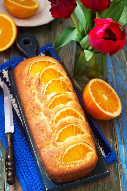 Orangenkuchen, verziert mit Orangenscheiben auf einem Hintergrund von saftigen Orangen und einem Strauß Tulpen. Rustikaler Stil.