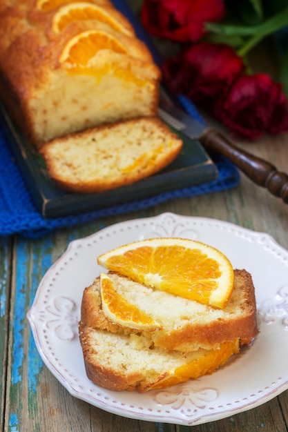 Orangenkuchen, verziert mit Orangenscheiben auf einem Hintergrund von saftigen Orangen und einem Strauß Tulpen. Rustikaler Stil.