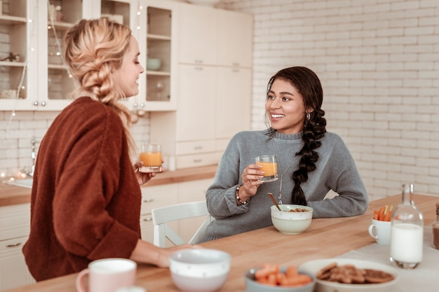 Orangensaft tragen. Angenehme freundliche Mädchen, die sich aktiv unterhalten, während sie sich auf den Tisch lehnen und einander ansehen