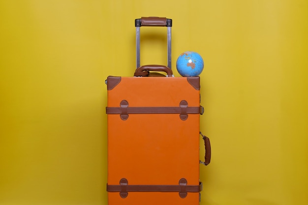 Oranger Koffer mit Mini-Globus isoliert auf gelbem Hintergrund für Reisekonzept mit minimalem Stil