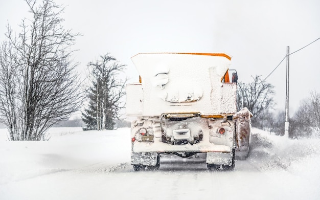 Oranger Pflug-LKW auf schneebedeckter Straße; grauer Himmel und Bäume im Hintergrund; Blick vom hinterfahrenden Auto - Winterdienst