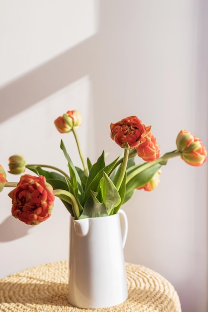 Orangerote Tulpenblumen mit grünen Stängeln und Blättern in einem weißen Keramikkrug auf einem hellen Schatten, weißer Wand- und Fensterhintergrund Kreative florale Botaniktapete Minimale kreative Grußkarte