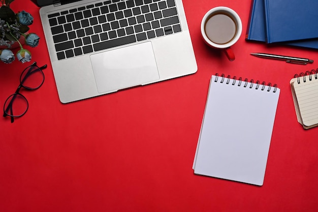 Ordenador portátil, cuaderno vacío, taza de café y vasos sobre fondo rojo.