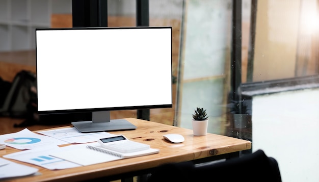 Ordenador portátil con pantalla en blanco en blanco en escritorio de madera que está rodeado de taza de café, pila de libros, planta en maceta, lápices sobre cómodas ventanas de la sala de estar como fondo.