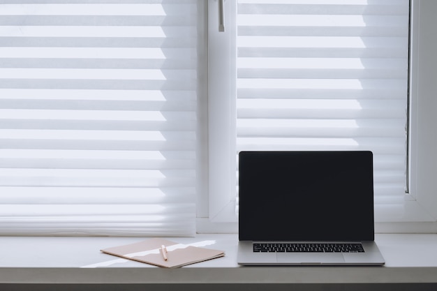 Ordenador portátil con pantalla en blanco y un cuaderno en la ventana en un día soleado. Hogar del lugar de trabajo