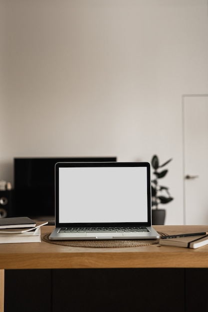 Ordenador portátil con pantalla en blanco, cuadernos de mesa de madera en la sala de luz solar cálida.