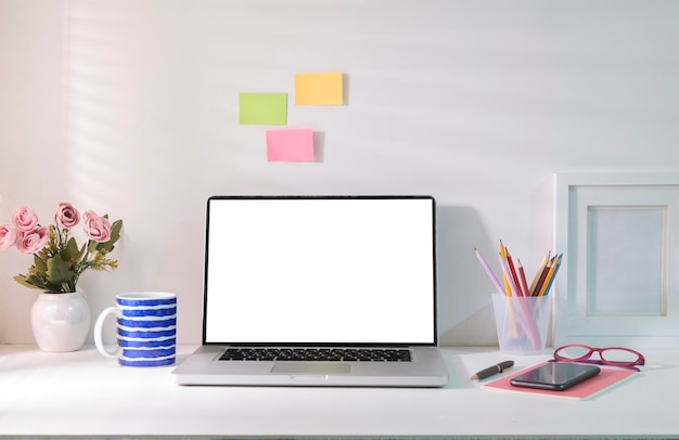 Ordenador portátil con pantalla en blanco, marco de fotos, maceta y cuaderno en mesa blanca