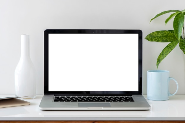 Ordenador portátil con pantalla en blanco en la mesa blanca con florero, libro, taza y planta de interior.