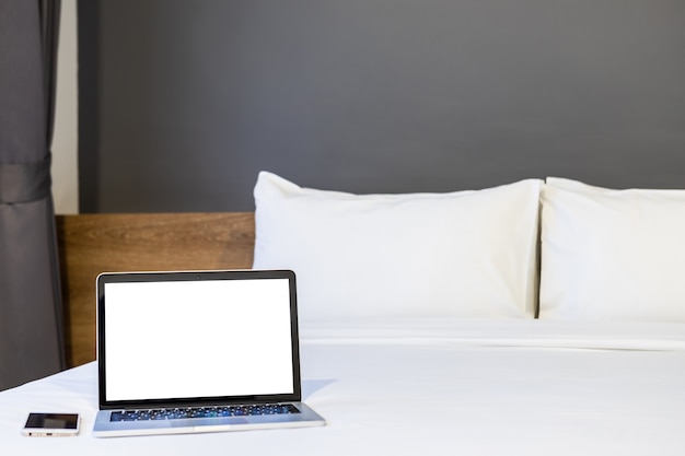 Ordenador portátil con pantalla en blanco y teléfono inteligente en la cama blanca en la habitación del hotel