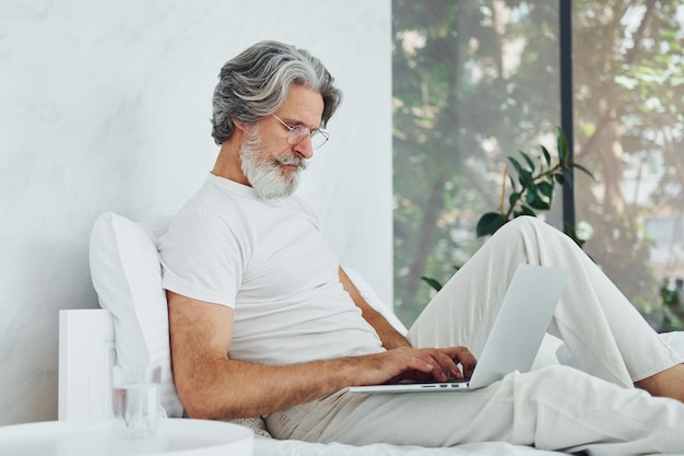 Ordenador portátil en las piernas Hombre moderno con estilo senior con cabello gris y barba en el interior