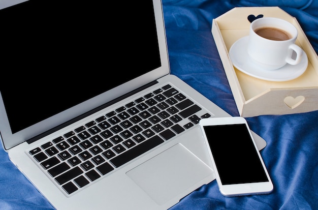 Ordenador portátil y teléfono inteligente, taza de café y cuaderno en la cama en el tiempo de la mañana. Bosquejo.