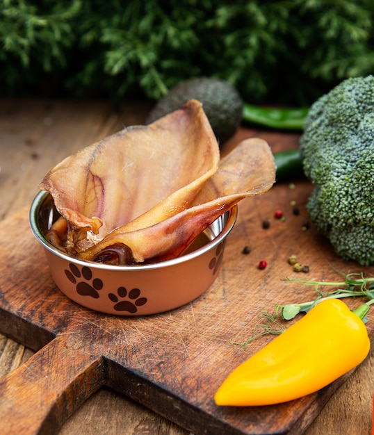 Orejas de cerdo secas en el recipiente del perro entre la vegetación en la tabla de madera. Masticar golosinas para perros domésticos.