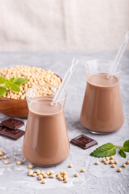 Organische nicht Molkereisojaschokoladenmilch in der Glas- und Holzplatte mit Sojabohnen auf einem grauen konkreten Hintergrund.