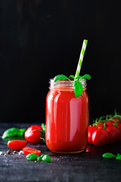 Organischer frischer Tomatensaft in einem Glasgefäß, in Basilikum, in Kirsche, in Salz, in Pfeffer und in Stroh auf dunklem Schwarzem. Sauberes Essen und Diätkonzept. Platz kopieren