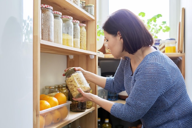 Organização de mulher de despensa na cozinha perto de rack de madeira com latas e recipientes de comida