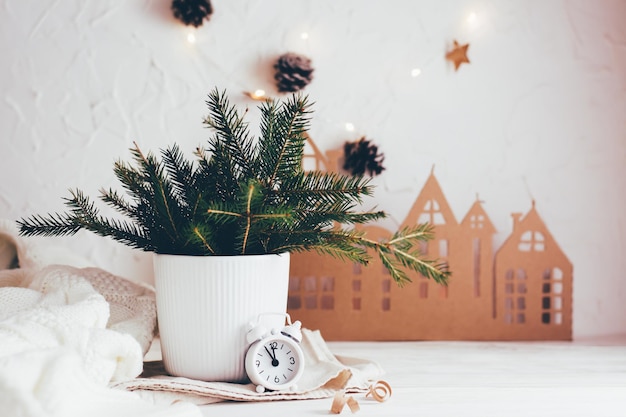 Organização de uma mesa festiva de Natal. Buquê de abetos em um vaso e um despertador branco no fundo de uma casa de papelão em uma mesa de madeira branca. Estilo de vida ecológico e caseiro