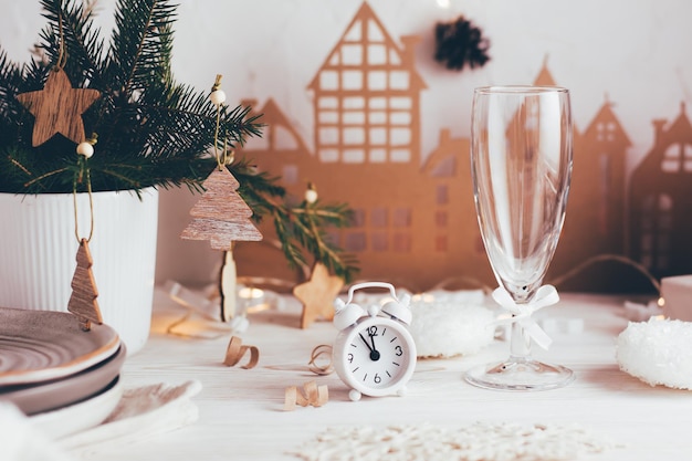 Organización de una mesa festiva navideña. Despertador blanco y una copa de champán en el fondo de un ramo de abeto en un jarrón y una casa de cartón. Paisajes de mesa en mesa de madera blanca