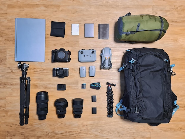 Organizar equipo de fotógrafo profesional con lente de cámara, mochila, estuche para computadora portátil, filtro, trípode, dron y saco de dormir en piso de madera