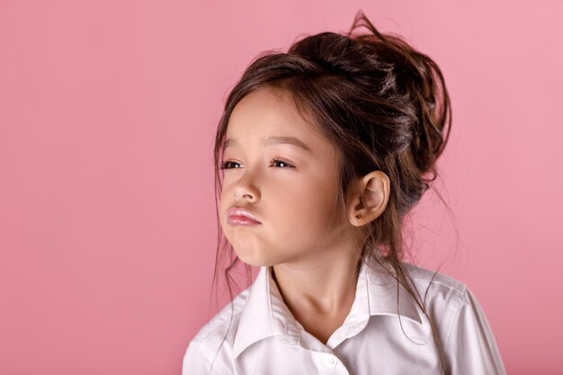 Orgulhosa garotinha na camisa branca com penteado, olhando para a câmera no fundo rosa. Eu sou o melhor. Emoções humanas