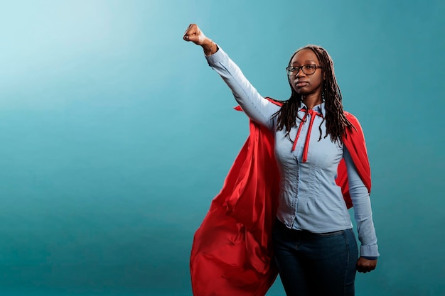 Orgulhoso e forte defensor da justiça afro-americana parecendo ambicioso enquanto olha para a câmera. Poderosa e corajosa jovem super-herói vestindo fantasia de herói enquanto posando como voando sobre fundo azul.