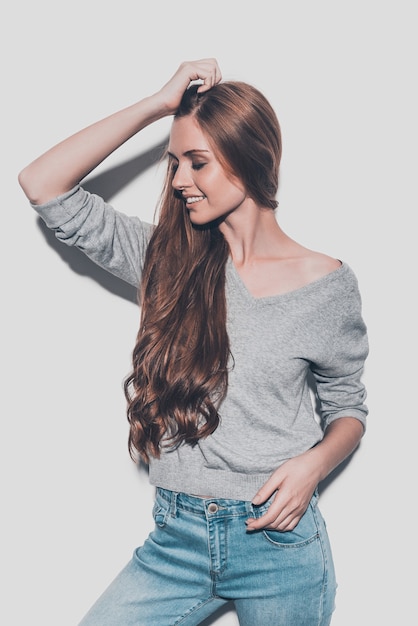 Foto orgullosa de su cabello sano. atractiva joven mujer sonriente sosteniendo la mano en el cabello y manteniendo los ojos cerrados mientras está de pie