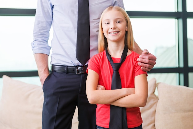 Orgullosa de su padre. Niña alegre en corbata de pie cerca de su padre y manteniendo los brazos cruzados