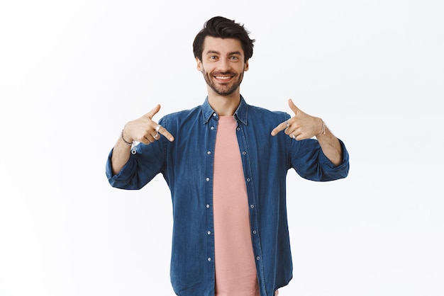 Orgulloso apuesto hombre caucásico barbudo confiado en ropa casual apuntando hacia abajo y sonriendo con expresión complacida y segura de sí misma invitando a visitar enlace anunciar producto dar consejos