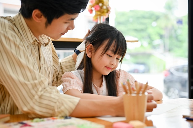 Orgulloso padre asiático acariciando la cabeza de su hija mientras le enseña la tarea en casa