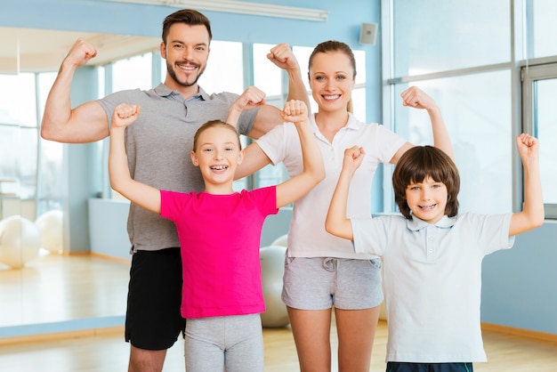 Orgulloso de ser fuerte y saludable. Familia deportiva feliz mostrando sus bíceps y sonriendo mientras está de pie cerca uno del otro en el club deportivo