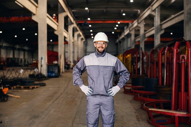 Un orgulloso trabajador de la industria pesada con ropa protectora se encuentra en la fábrica y sonríe a la cámara