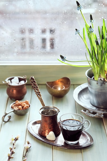 Orientalischer Kaffee in traditioneller türkischer Kupferkaffeekanne mit Blumen auf der Fensterbank Holzfensterbank mit Hyazinthenblumentopf Kalter Regentag im Frühling Gemütliches Szene-Hygge-Konzept