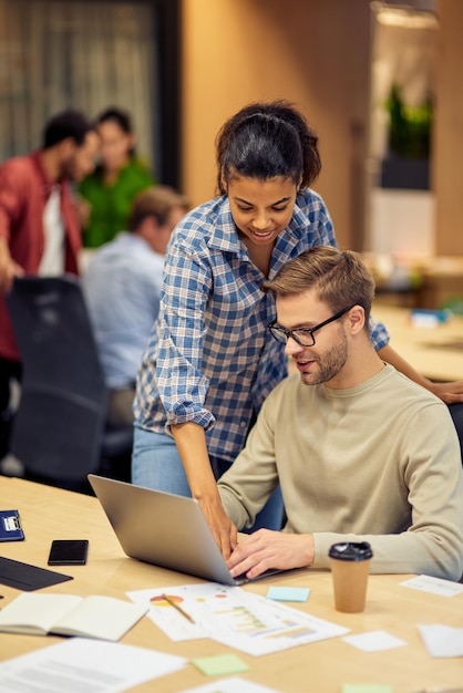 Orientando no escritório dois jovens colegas multirraciais usando laptop e discutindo projeto de raça mista