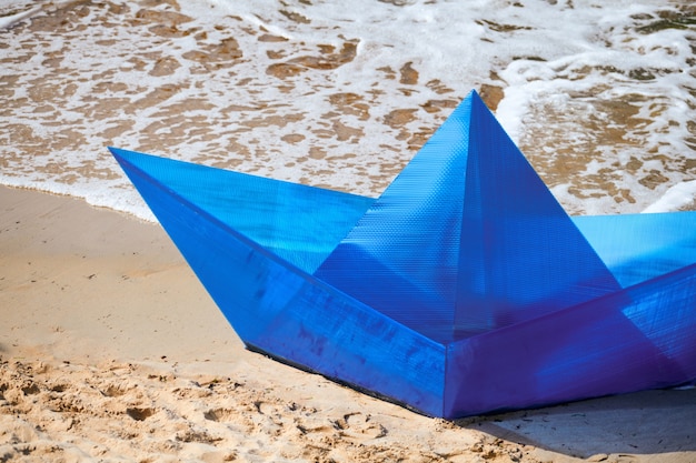 Origami blaues Papierboot am Sandstrand für Konzeptdesign, schönes blaues Papierschiff am Ufer. Objekt der modernen Kunst, Nahaufnahme, Meereshintergrund. Reise- und Sommerurlaub, Träumen und Vorstellungskonzept