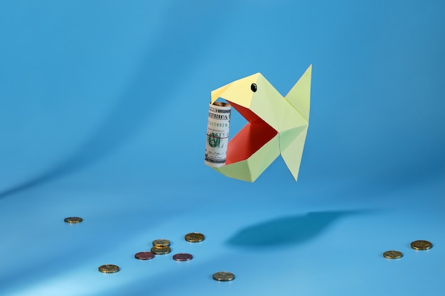 Origami-Fisch in offenem Mund hält sich in Röhrendollar auf dem Hintergrund kleiner Münzen unten verdreht