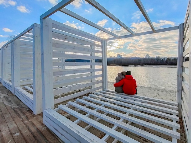 En la orilla de un río de invierno, un hombre y una mujer con chaquetas están sentados en una estructura decorativa