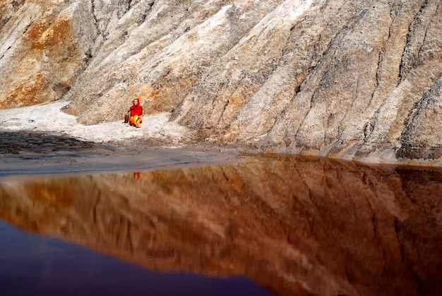 Orilla rocosa de un lago en el desierto y la figura solitaria de una mujer sentada en tierra con una jarra