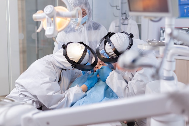 Ortodentista y asistente que trata a una niña con instrumentos modernos vestida con traje de ppe. Equipo de estomatología con traje de ppe durante el procedimiento covid19 en dientes infantiles.