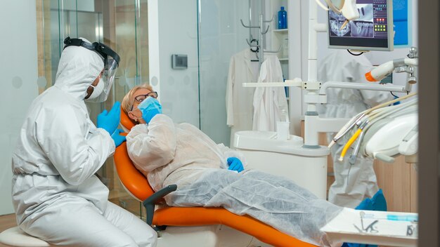Ortodoncista en mono de protección hablando con paciente con dolor de muelas durante covid-19 en consultorio estomatológico. Concepto de nueva visita al dentista normal en el brote de coronavirus con traje protector y