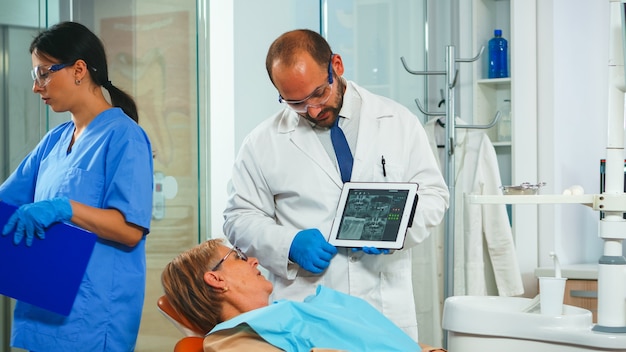 Ortodontista usando tablet explicando o raio-x odontológico ao paciente sentado na cadeira odontológica no consultório de estomatologia. dentista, mostrando a radiografia de velha usando dispositivo digital, trabalhando na clínica moderna.